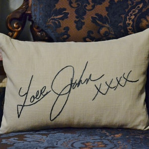 Puede incluir: Una almohada beige con una firma bordada en negro que dice "Love Josh xxx" en un sof&aacute; de estampado azul oscuro y marr&oacute;n.