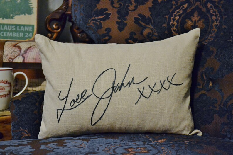 Puede incluir: Una almohada beige con una firma bordada en negro que dice "Love Josh xxx" en un sof&aacute; de estampado azul oscuro y marr&oacute;n.