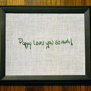 May include: A black framed picture with a white fabric background. The fabric has green embroidery that reads "Poppy loves you so much!"