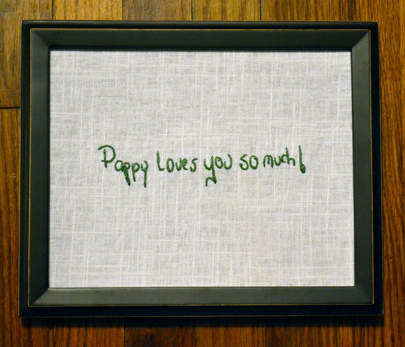 May include: A black framed picture with a white fabric background. The fabric has green embroidery that reads "Poppy loves you so much!"