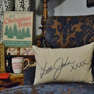 Puede incluir: Un letrero de estilo vintage que anuncia una granja de &aacute;rboles de Navidad con el texto "KRINGLE FAMILY TREE FARM CHOOSE Christmas Trees 4301 SANTA CLAUS LANE OPEN THRU DECEMBER 24". Un coj&iacute;n blanco con el texto "Love John XXXX" est&aacute; en un sof&aacute; de estampado marr&oacute;n y azul.