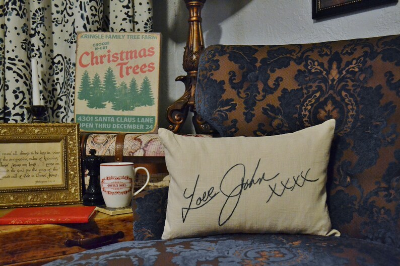 Puede incluir: Un letrero de estilo vintage que anuncia una granja de &aacute;rboles de Navidad con el texto "KRINGLE FAMILY TREE FARM CHOOSE Christmas Trees 4301 SANTA CLAUS LANE OPEN THRU DECEMBER 24". Un coj&iacute;n blanco con el texto "Love John XXXX" est&aacute; en un sof&aacute; de estampado marr&oacute;n y azul.