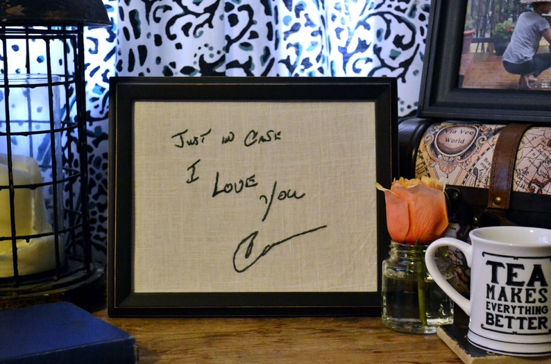 May include: A framed message on a beige fabric background reads "Just in case I love you" with a handwritten signature. The frame is black with a brown border. The message is on a table with a rose in a glass jar and a white mug with the text "Tea makes everything better".