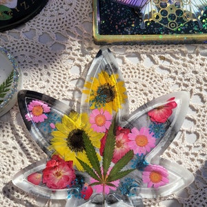 Puede incluir: Una bandeja de resina transparente con forma de hoja de marihuana con flores y vegetación prensadas en su interior. Las flores son rosas, amarillas y blancas. La vegetación es de color verde oscuro.