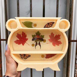 Pode incluir: Um caldeirão de cerâmica cor de creme com três níveis, com flores, folhas e borboletas secas prensadas. O caldeirão tem uma base arredondada e uma parte superior larga e aberta.