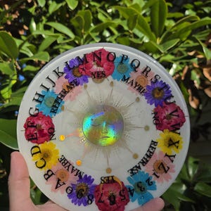 Puede incluir: Una tabla Ouija de resina blanca con flores secas coloridas y detalles dorados. La tabla presenta el alfabeto, números y las palabras "SÍ" y "QUIZÁS". Un diseño de sol holográfico está en el centro.