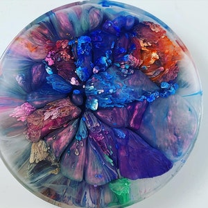 May include: A round, resin coaster with an abstract design. The coaster features a blend of colors, including blue, purple, pink, and orange, creating a swirling, artistic pattern. The edges are clear, and the overall effect is glossy.