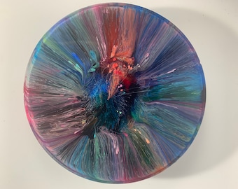 May include: A round, abstract resin art piece with a vibrant color palette. The artwork features a central burst of red, orange, and pink hues, surrounded by flowing lines of blue, green, and purple. The piece has a glossy finish.