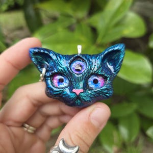 May include: A blue cat pendant with three eyes, a pink nose, and large blue and pink eyes. The cat pendant has a silver bail and a small silver hoop on one ear. The pendant is held in a hand.