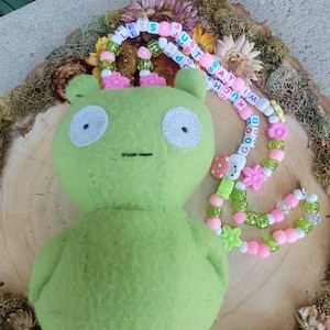 以下が含まれることがあります： 大きな目とシンプルな口をした緑色のぬいぐるみ。おもちゃは、木製の表面に置かれ、ピンクと緑のビーズネックレスがかけられています。ビーズには文字が書かれており、「LIFE IS JUST A DREAM」と「GOOD GOOD GOOD」と綴られています。