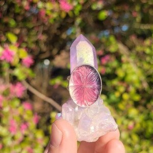 May include: A silver ring with a pink and purple flower design set in a clear glass cabochon. The ring is displayed on a white crystal.