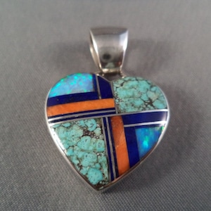 Könnte beinhalten: Ein silberner herzförmiger Anhänger mit Türkis-, Lapislazuli- und Opal-Einlegearbeiten. Der Anhänger hat ein geometrisches Design mit einem Kreuzmuster.