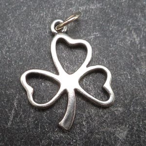 以下が含まれることがあります： シルバーカラーの三つ葉のクローバーペンダント。チェーンを通す小さなループが付いています。幸運の象徴である三つ葉のクローバーのデザイン。暗い背景に配置されています。
