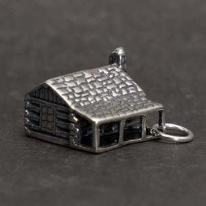 May include: A silver charm in the shape of a log cabin with a chimney and a small window. The charm has a loop for attaching to a bracelet or necklace.