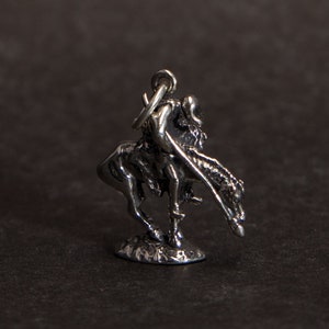 May include: Silver charm depicting a Native American on horseback. The charm is detailed and features a rider with a spear and a horse with a flowing mane.