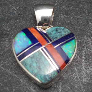 Puede incluir: Un colgante de plata en forma de corazón con un diseño geométrico hecho de piedras turquesa, azules, rojas y ópalos.