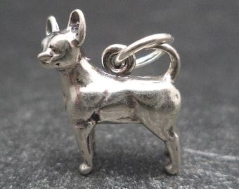 May include: Silver charm of a Chihuahua dog standing on all four legs. The charm has a loop at the top for attaching to a bracelet or necklace.