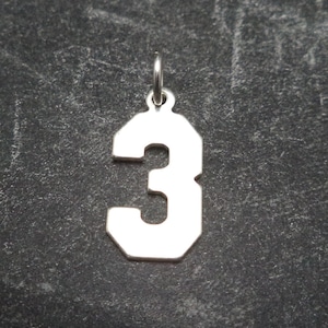 May include: A silver charm in the shape of the number 3. The charm is a simple, solid design with a slightly rounded edge.