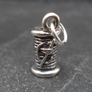 May include: Silver charm in the shape of a spool of thread with a needle. The charm has a loop for attaching to a bracelet or necklace.