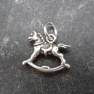 May include: A silver rocking horse charm. The horse is in a rocking motion with its legs raised. The charm has a loop at the top for attaching to a necklace or bracelet.