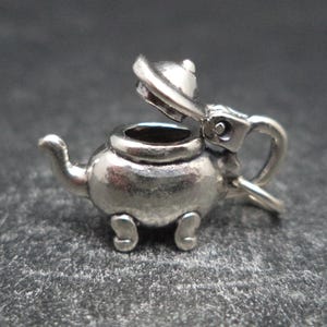 May include: A silver charm in the shape of a teapot with the lid open. The teapot has a handle and a spout and is standing on four small feet.