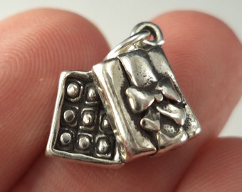 May include: Silver charm in the shape of a closed book with a heart design on the front cover. The charm is detailed with a textured surface and a loop for attaching to a bracelet or necklace.