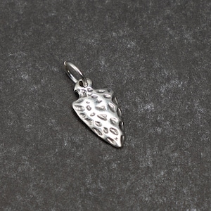 May include: A silver arrowhead charm with a textured surface. The charm has a loop at the top for attaching to a necklace or bracelet.