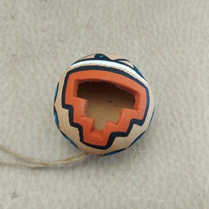 Puede incluir: Un pequeño adorno redondo de terracota con un diseño geométrico en naranja, azul y blanco. El adorno tiene un agujero en la parte superior y una cuerda atada para colgarlo.