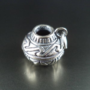 Puede incluir: Un charm de plata con forma de olla pequeña, con detalles intrincados y un diseño decorativo. El charm tiene un bucle para sujetarlo a una pulsera o collar.