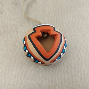 Puede incluir: Una pequeña maceta redonda de terracota con un diseño geométrico pintado en naranja, negro, blanco y azul. La maceta tiene un agujero en la parte superior y una cuerda atada para colgarla.