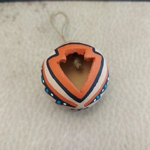 Puede incluir: Una pequeña maceta de arcilla redonda, naranja y marrón con un diseño geométrico. La maceta tiene un agujero en la parte superior y una cuerda atada para colgarla.