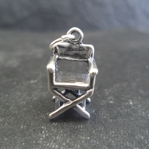 May include: Silver charm in the shape of a director's chair. The chair has a simple design with a back, seat, and four legs.