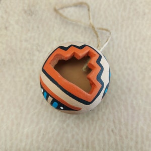 Puede incluir: Un pequeño adorno redondo de terracota con un diseño geométrico en naranja, negro y azul. El adorno tiene un agujero en el centro y una cuerda atada para colgarlo.