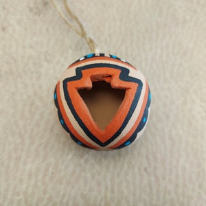 Puede incluir: Un pequeño adorno redondo de arcilla de color naranja, negro y blanco con un diseño geométrico. El adorno tiene un agujero en el centro y una cuerda atada para colgarlo.