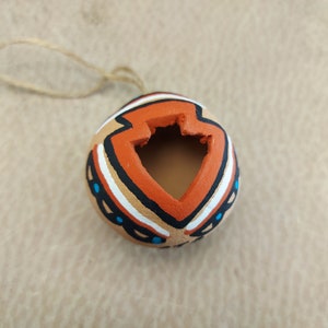 Puede incluir: Un pequeño adorno redondo de arcilla de color naranja, marrón, blanco y azul con un diseño geométrico. El adorno tiene un agujero en la parte superior para colgarlo.