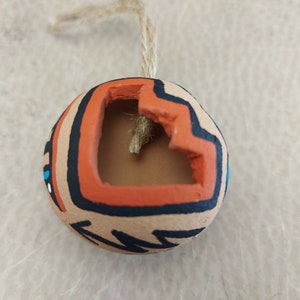 Puede incluir: Un pequeño adorno redondo de terracota con un agujero en la parte superior para colgar. El adorno está decorado con patrones geométricos negros, naranjas y azules.
