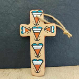 Puede incluir: Un adorno de cruz de terracota con un diseño geométrico que presenta triángulos naranjas, blancos y azules. La cruz tiene una cuerda para colgar.
