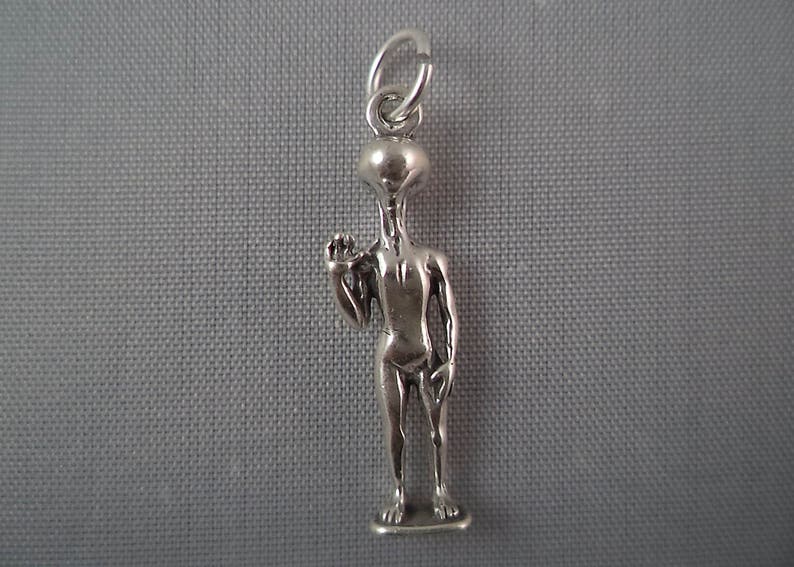 May include: A silver charm in the shape of a humanoid alien with large eyes and a raised hand. The charm is attached to a silver loop.