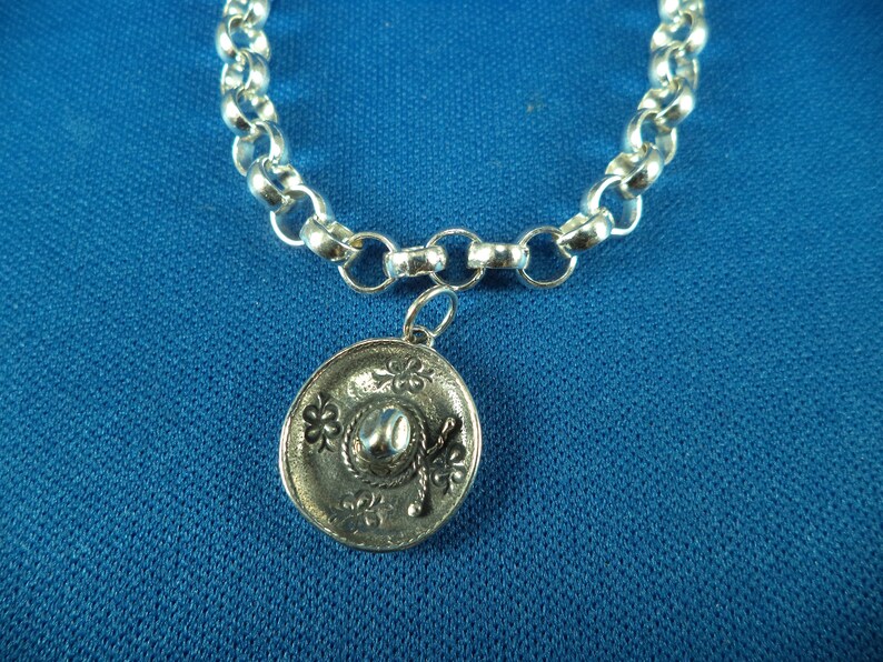 May include: A silver chain bracelet with a silver charm in the shape of a sombrero. The charm has a detailed design with a flower pattern.