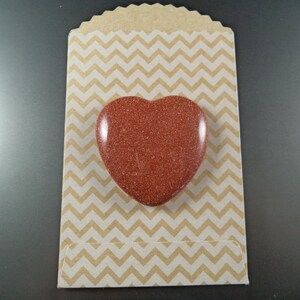 May include: A heart-shaped red stone with a glittery finish, displayed on a brown and white chevron patterned paper bag.