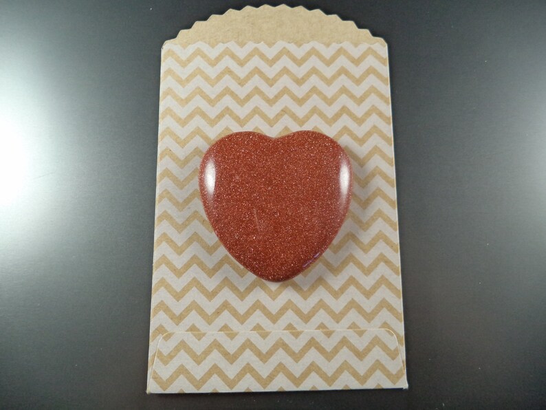 May include: A heart-shaped red stone with a glittery finish, displayed on a brown and white chevron patterned paper bag.
