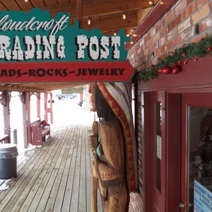Op de afbeelding: Een houten bord met de tekst "Cloudcroft Trading Post" en de woorden "Beads-Rocks-Jewelry" eronder. Het bord is boven een houten veranda met een rode bakstenen muur aan de rechterkant gemonteerd. Een houten figuur van een indiaan staat voor de bakstenen muur.