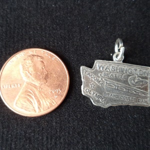 Op de afbeelding: Zilveren bedel in de vorm van de staat Washington met de steden Seattle, Tacoma en Olympia, en de tekst "Grand Coulee Dam" en "Washington" erop. De bedel ligt naast een Amerikaanse penny ter groottevergelijking.