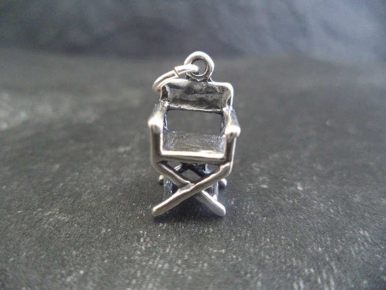 May include: Silver charm in the shape of a director's chair. The chair has a simple design with a back, seat, and four legs.