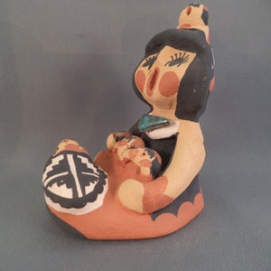 May include: A hand-painted ceramic figurine of a Native American woman holding a baby. The woman is wearing a black dress with a turquoise stone and has black hair. The figurine is painted in shades of brown, black, white, and turquoise.