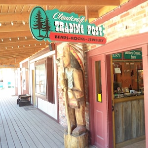 May include: A wooden sign with the text "Cloudcraft Trading Post Beads-Rocks-Jewelry" hangs above a wooden door with a carved wooden Native American figure standing next to it.