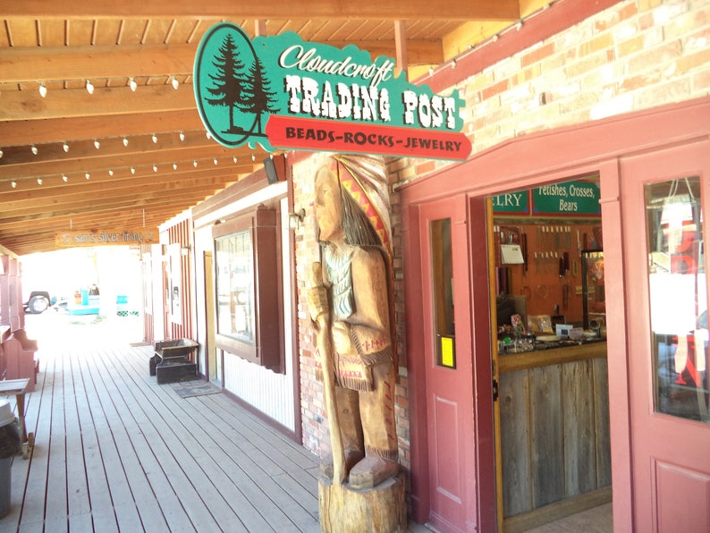 May include: A wooden sign with the text "Cloudcraft Trading Post Beads-Rocks-Jewelry" hangs above a wooden door with a carved wooden Native American figure standing next to it.