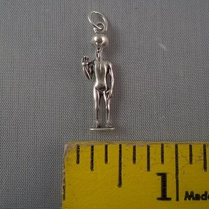 May include: Silver charm of a gray alien with a large head and thin body. The alien is standing with one arm raised and a finger pointing up. The charm is about 1 inch tall and is shown next to a ruler.