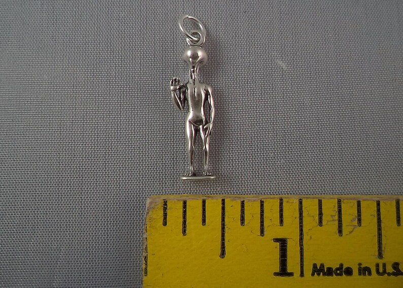 May include: Silver charm of a gray alien with a large head and thin body. The alien is standing with one arm raised and a finger pointing up. The charm is about 1 inch tall and is shown next to a ruler.