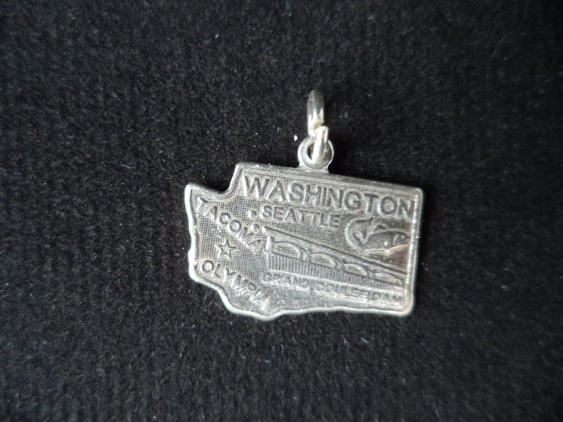 Op de afbeelding: Zilveren bedel in de vorm van de staat Washington met de woorden "WASHINGTON", "SEATTLE", "TACOMA", "OLYMPIA" en "GRAND COULEE DAM" erop.
